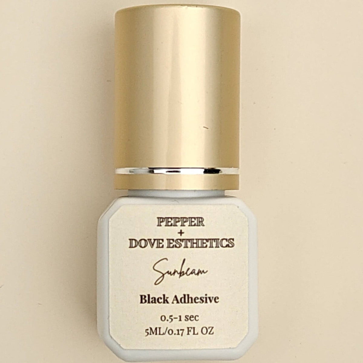 A bottle of Pepper + Dove Esthetics Selen Black Adhesive with a metallic gold cap and a white label, centered on a warm beige background.