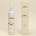 A white bottle of Pepper Dove Botanicals Whipped Clean Cleanser stands next to its cream-colored rectangular box on a warm beige background.