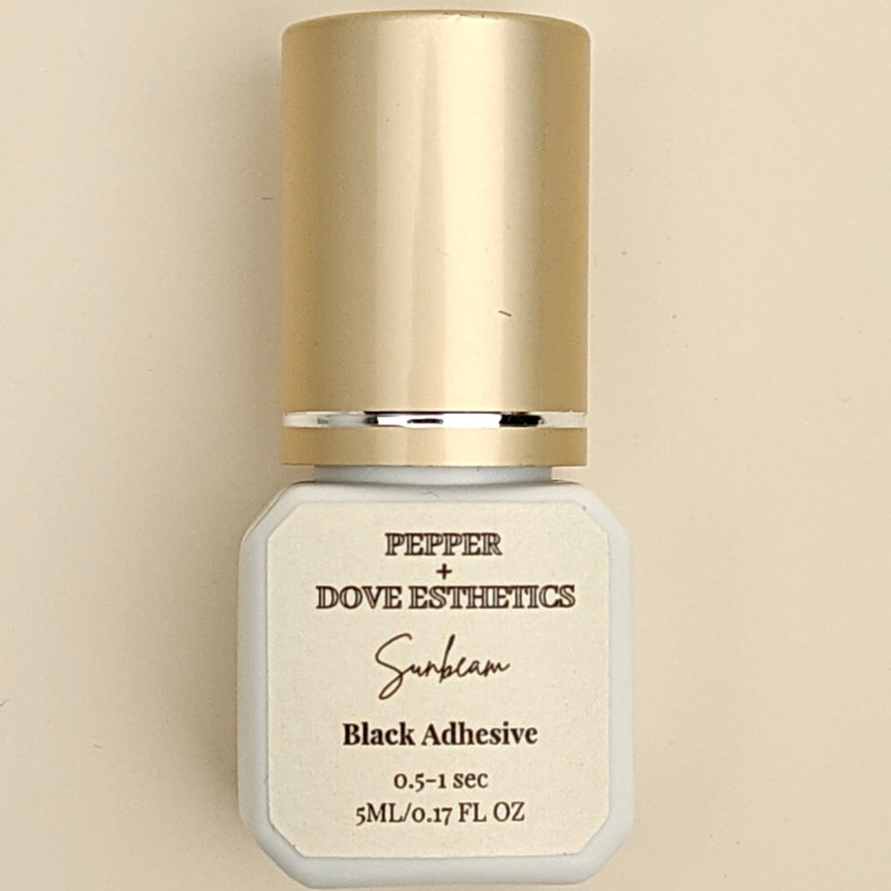 A bottle of Pepper + Dove Esthetics Selen Black Adhesive with a metallic gold cap and a white label, centered on a warm beige background.