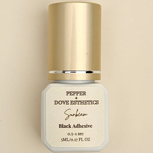 A bottle of Pepper + Dove Esthetics Selen Black Adhesive with a metallic gold cap and a white label, centered on a warm beige background.